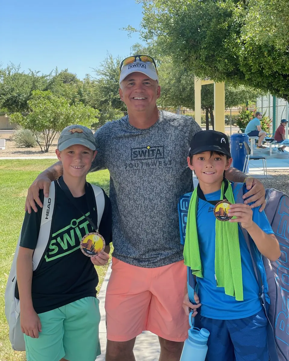 Two young tennis players stand proudly with their coach in a sunny park, holding medals from a recent competition.